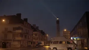 Een foto van een straat in een Franse stad. Het is donker. Op de achtergrond zie je een vuurtoren die een lichtstraal uitzendt. Je ziet geen mensen op straat lopen. 