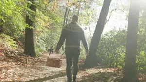 Een foto van iemand die over een pad door het bos loopt met een mand in zijn hand. Het is erg zonnig en het zonlicht schijnt tussen de bomen.