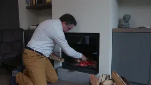Een foto van een man die in de huiskamer de haard aan het vullen is met hout. In de haard zijn kolen te zien die rood gloeien van de hitte.