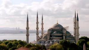 Een foto van de islamitische Süleymaniye-moskee in Istanbul. Te zien zijn vijf torens van lichtbruine tot grijze stenen met goudkleurig versierde punten en blauwgrijze, ronde daken. De grote koepel bovenop heeft een goudkleurige halve maan als punt.