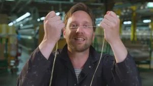 Een foto van presentator Bart die in een dradenfabriek staat. Hij houdt een sterke draad tussen zijn handen en voor zijn gezicht en trekt er hard aan. Hij kijkt in de camera. 