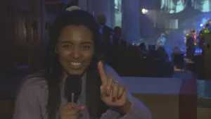 Een foto van presentator Sosha die voor de camera staat in het publiek van een gospel optreden. Gospel betekent 'Goede boodschap' en is een soort muziek.