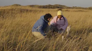 Een foto van Sosha en Sorosh die op hun knieën zitten op een veld vol gele en groene grashalmen. Er steekt een ronde, witte staaf uit de grond, een sensor die de bodemvochtigheid meet. Aan een grijze staaf zit een wit kastje om de metingen door te geven.