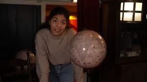 Een foto van een presentator die de camera inkijkt. Voor haar staat er een wereldbol. 