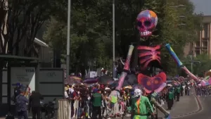 Een foto van een optocht bij de viering van de Dag van de Doden in Mexico. Er is een grote, kleurrijke pop van een skelet te zien en iemand die ervoor loopt met groene en oranje kleding en een grijs doodshoofd masker. Mensen achter hekken kijken ernaar.