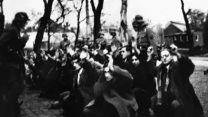 Een zwart-wit foto genomen tijdens de Februaristaking in 1941. Er zitten mensen op de grond met hun handen omhoog. Ze kwamen in verzet tegen de Jodenvervolging door nazi-Duitsland. Er staan soldaten met helm, links één met geweer en vier achteraan.