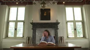 Een foto van Pascal die achter een rechthoekige, bruine tafel zit. Voor hem op tafel ligt een dik, opengeslagen boek met aan de zijkant bruingekleurde bladzijden. Aan de muur hangt een portret van Constantijn Huygens, de vader van Christiaan Huygens.