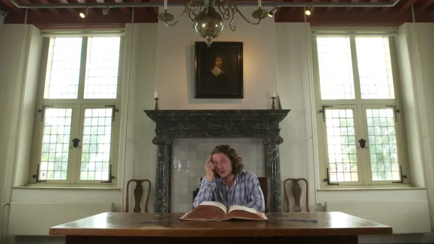 Een foto van Pascal die achter een rechthoekige, bruine tafel zit. Voor hem op tafel ligt een dik, opengeslagen boek met aan de zijkant bruingekleurde bladzijden. Aan de muur hangt een portret van Constantijn Huygens, de vader van Christiaan Huygens.