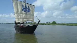 Een foto van een schip van zwartgekleurd hout in het water. Het schip heeft één groot wit zeil, waarop in een blauwe kleur een kasteel staat met drie torens. Er zijn van veraf drie zeemannen op het schip te zien in rode jas en met een zwarte hoed. 