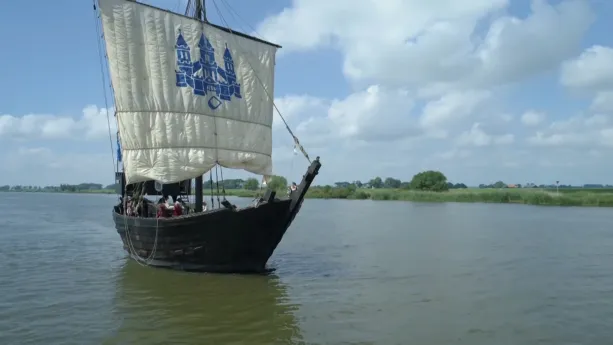 Een foto van een schip van zwartgekleurd hout in het water. Het schip heeft één groot wit zeil, waarop in een blauwe kleur een kasteel staat met drie torens. Er zijn van veraf drie zeemannen op het schip te zien in rode jas en met een zwarte hoed.