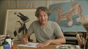 Een foto van een tekenaar die in de camera kijkt. Hij zit aan tafel en voor hem ligt er een vel papier waarop hij iets aan het tekenen is. Achter hem staan schilderijen van twee dieren.