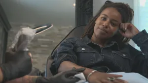 Een foto van presentator Eva. Ze zit in een stoel en heeft haar rechterarm ontbloot op een tafel liggen. Ze kijkt in de camera. Een tatoeëerder wrijft met iets over haar arm. 