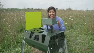 Een foto van presentator Manon die buiten naast een insecten camera staat waar het aantal en soort insecten mee geteld wordt.