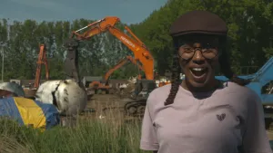 Een foto van presentator Eva. Ze staat op het terrein van een scheepssloperij en kijkt enthousiast in de camera. Op de achtergrond zie je diverse graafmachines en grote ijzeren onderdelen.
