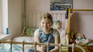 Een foto van de negenjarige Levin. Ze zit op haar bed en kijkt naar iets buiten beeld. Op de achtergrond zie je wat knuffels, en een schilderij aan de muur.