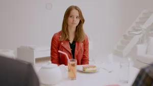 Een foto van een tienermeisje. Ze zit aan een tafel en kijkt naar iets buiten beeld. Voor haar ligt een tompouce op een bord, en een glas limonade. Op de achtergrond zie je een witte trap.