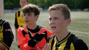 Een foto van twee jongens in voetbalkleren. Ze staan net buiten het veld en kijken in dezelfde richting. Ze zijn in gesprek met iemand en ze kijken daarbij ernstig.