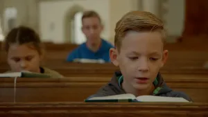 Een foto van een jongen die op een houten kerkbank zit. Voor zich heeft hij een bijbel opengeklapt liggen, waaruit hij lees of zingt. Erachter zit een meisje en nog verder naar achteren een man op een houten kerkbank die ook een bijbel voor zich hebben.