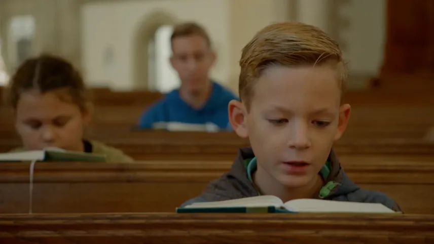 Een foto van een jongen die op een houten kerkbank zit. Voor zich heeft hij een bijbel opengeklapt liggen, waaruit hij lees of zingt. Erachter zit een meisje en nog verder naar achteren een man op een houten kerkbank die ook een bijbel voor zich hebben.