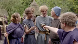 Een foto van een interviewer met een soort microfoon en vier andere mensen: een vrouw in een blauw kostuum met een stok in handen, een man in een grijs kostuum, een man in een lichtgrijs kostuum en  een man met een bruin kostuum en een blauwgrijze hoed.