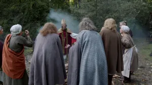 Een foto van een man verkleed als Bonifatius in een rood pak vergelijkbaar met dat van Sinterklaas met een goudkleurige meiter op zijn hoofd, een goudkleurige cape en een goudkleurige sjaal om. Er staat een groep mensen in een kring om hem heen. 