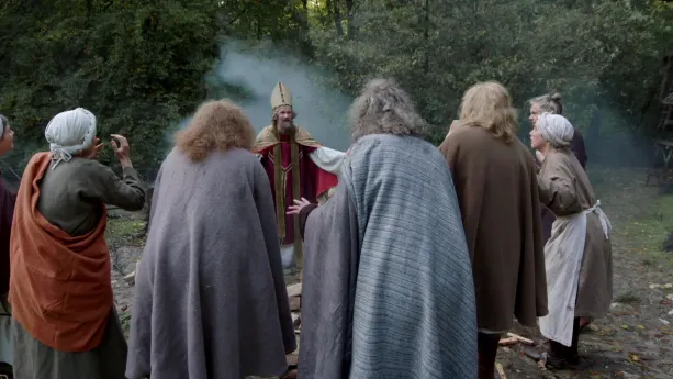 Een foto van een man verkleed als Bonifatius in een rood pak vergelijkbaar met dat van Sinterklaas met een goudkleurige meiter op zijn hoofd, een goudkleurige cape en een goudkleurige sjaal om. Er staat een groep mensen in een kring om hem heen.