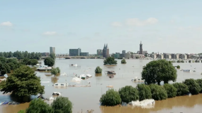 Een foto van een bewoond gebied achteraan met gebouwen, woningen en een brug. Ervoor ligt een grote vlakte van hoog water. Er liggen meer dan tien weggedreven caravans en stacaravans in het water en er staan bomen tot aan de groene kruin in het water.