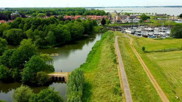 Een foto van een smalle, geasfalteerde weg over een dijk, een groene heuvel die langs het water naar beneden loopt. Erachter liggen boten in het water. Aan de andere kant van het water staan een molen en huizen.