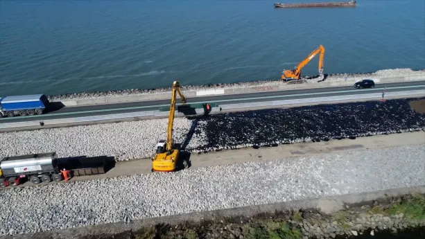Een foto van twee graafmachines: één direct naast het water, één naast een weg met een auto en vrachtauto. Er liggen lichtgrijze stenen naast, die een dijk vormen. Door de graafmachines zijn de stenen al deels voorzien van een zwarte, verstevigende laag.