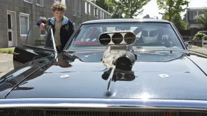 Een foto van een jongen die een grote auto instapt aan de passagierskant en kijkt naar de motorkap waar de motor op ligt.