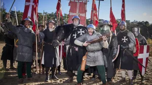 Een foto van een groep Middeleeuwse ridders die allemaal richting de camera kijken. Sommigen van hen schreeuwen. Ze hebben wapens bij zich, en een paard. Op de achtergrond zie je rode vlaggen met daarop witte kruizen.