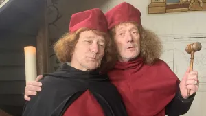 Een foto van twee mannen in Middeleeuwse kleren. De een heeft zijn arm om de schouder van de ander. Hij heeft een houten hamer in zijn handen. Op de achtergrond zie je een grote brandende kaars.