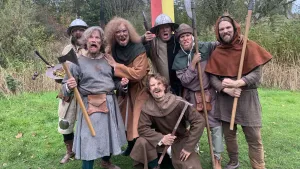 Een foto van zeven mannen in Middeleeuwse kleren. Ze staan buiten, op het gras, en schreeuwen in de camera. Ze hebben bijlen, speren en andere wapens bij zich. 