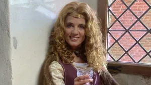 Een foto van een vrouw met de Middeleeuwse kleren. Ze zit in een vensterbank en houdt een glas vast. Ze heeft lange blonde krullende haren en kijkt in de camera.