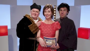 Een foto van een vrouw en twee mannen in een Tv-studio. De vrouw heeft moderne kleren aan. De mannen hebben Middeleeuwse kleren aan. De vrouw is de presentator en ze houdt een kaartje vast met daarop de tekst 'Welkom in de Middeleeuwen'.