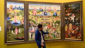 Een foto van een presentator die naar de rechterkant van het schilderij "De Tuin der Lusten" wijst, waar twee schuin gedraaide schilderijen aan vastzitten. Er staan mensen op en op twee delen veel licht, groen en water. Het rechterdeel is erg donker. 