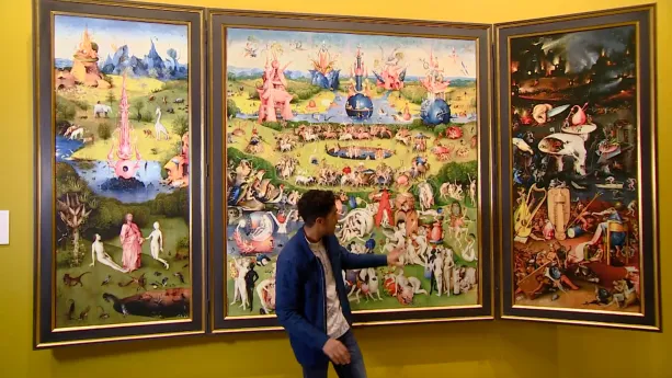 Een foto van een presentator die naar de rechterkant van het schilderij "De Tuin der Lusten" wijst, waar twee schuin gedraaide schilderijen aan vastzitten. Er staan mensen op en op twee delen veel licht, groen en water. Het rechterdeel is erg donker.