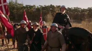 Een foto van soldaten op een zandvlakte in het bos. Ze dragen een metalen helm, een maliënkolder, een kledingstuk van metalen ringetjes en vier rode vlaggen met een wit kruis erop. Een man op een bruin paard draagt zwarte kleding met een wit kruis erop.