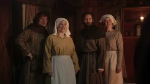 Een foto van vier mensen die naar iets rechtsachter de camera kijken. De twee mannen dragen een bruin kostuum met zwarte rand om hun nek en schouders. De twee vrouwen dragen een lichtbruine trui, blauwe en lichtbruine rok en een witte doek om hun hoofd.