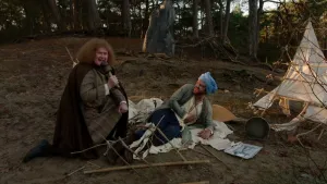 Een foto van een interviewer en een man verkleed als Ibn Firnas die op een witte doek ligt met een blauwe doek, een tulband, om zijn hoofd en zijn rechterhand op zijn buik. Een rek van stokkken ligt op zijn benen en zit ook aan een tent erachter vast. 