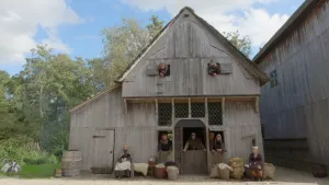 Een foto van een grijskleurig houten huis zoals mensen in de middeleeuwen hadden. Uit raamkozijnen met luiken, twee boven en twee beneden en het gat bovenin de deur steekt een persoon in ouderwetse kleding. Voor het huis zit links en rechts ook iemand.