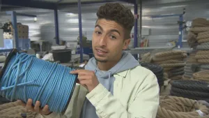 Een foto van presentator Nizar in een touwenfabriek. Hij houdt een grote klos vast. Daaromheen zit een blauw touw gewikkeld. Op de achtergrond zie je allerlei hele dikke touwen.