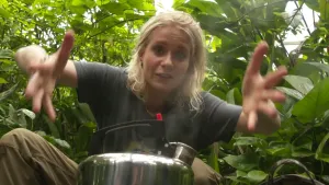 Een foto van Janouk die achter een stalen fluitketel zit in een kas met groene planten. Een fluitketel is een ketel om water mee te koken en als het kookt, komt uit de ronde opening een fluitend geluid. Janouk heeft haar handen wijd uit elkaar.