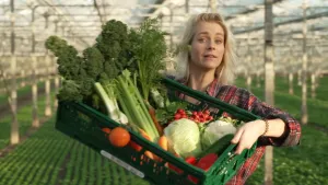 Een foto van Janouk die in een kas staat. Ze houdt een groene plastic bak omhoog met allemaal soorten groenten erin. In de bak liggen de volgende groenten: bloemkool, sla, boerenkool, radijsjes, tomaten, paprika, wortels en komkommer.