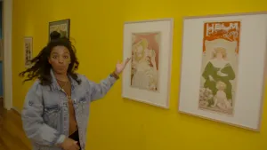 Een foto van Sosha die met haar hand wijst naar twee witte schilderijlijsten op een gele muur. In de lijsten zitten tekeningen: één van een vrouw die een kind een lepel cacao of chocolade van het merk Helm geeft, één van een naar beneden kijkende vrouw.