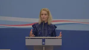 Een foto van Janouk achter een hoge spreektafel met een zwarte microfoon. Ze houdt haar handpalmen omhoog en draagt een blauwe blouse met witte strepen. Het Rijksoverheid logo staat op de tafel: twee leeuwen, ertussen een schild en erboven een kroon. 