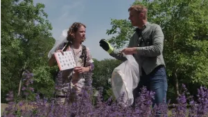 Een foto van Pascal en een man achter een rij paarse bloemen. Pascal heeft een papier met plaatjes vast, niet goed te zien, maar het zijn bijensoorten. De man kijkt ernaar en heeft een wit net met ronde opening vast en een gehandschoende hand omhoog.