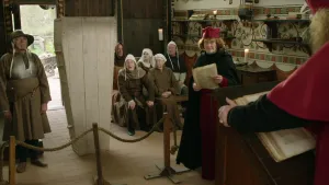 Een foto van een zaal met mensen die zitten op een houten bank. Vóór de mensen staat een houten doodskist rechtop, ernaast een soldaat met helm. Een man in rood pak, hoed en zwarte cape met een papier in handen, kijkt naar een man achter een hoge tafel.