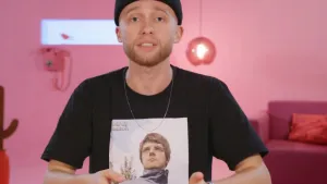 Een foto van presentator Jard. Hij kijkt in de camera. Op zijn T-shirt staat een foto van een man met een bril. De achtergrond bestaat uit een roze muur met een vaste telefoon daaraan, en een roze lamp en een roze bankstel.