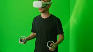 Een foto van presentator Jard. Hij staat in een ruimte met groene wanden. Hij heeft een Virtual Reality bril op, en in zijn handen heeft hij controllers vast. 